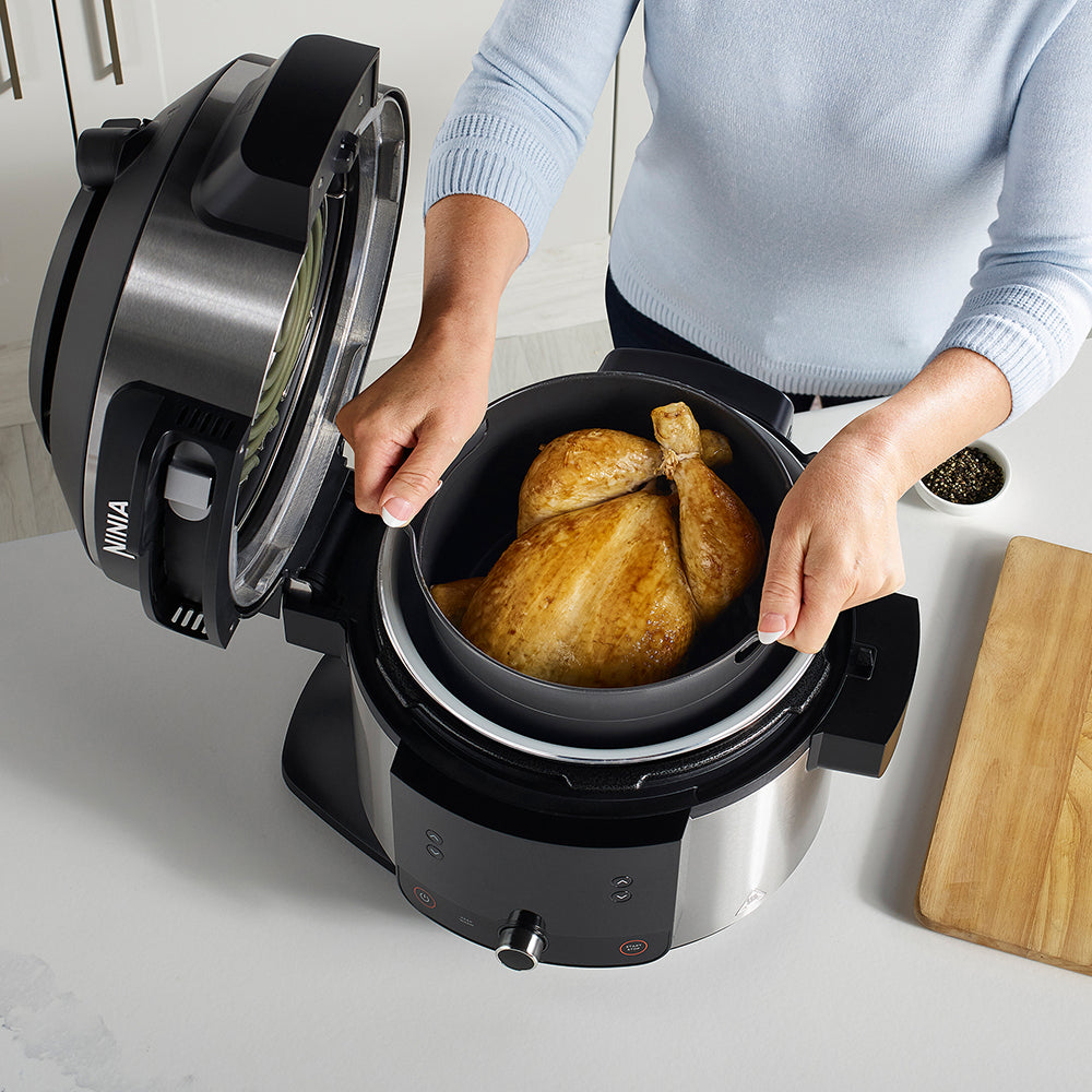 Person opening a Ninja foodi pressure cooker with roasted chicken inside on a kitchen counter.